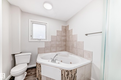 Bathroom featuring a corner jetted tub with tile surround, a built-in toilet, a window, and wood-finish flooring - 8615 42A Avenue, Edmonton, AB - Indoor Photo Showing Bathroom