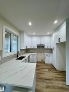 Kitchen featuring white shaker-style cabinetry, quartz countertops with a waterfall edge, a double basin sink with a matte black faucet, and wood-finish flooring - 
