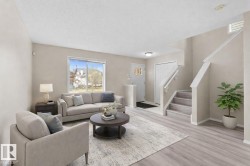 Open-concept living area featuring light wood-finish flooring, a contemporary staircase with white risers and handrails, and a front entry door with decorative glass inserts - 