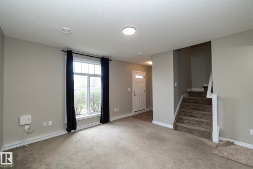 Open concept living space featuring neutral gray walls, plush carpet flooring, a multi-pane window with black drapery, and a visible staircase with carpeting - 117 Callaghan Drive Sw, Edmonton, AB - Indoor Photo Showing Other Room