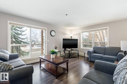 The living room features dark wood flooring, light grey walls, and ample natural light from a large window and sliding glass door - 202 17511 98A Avenue, Edmonton, AB - Indoor Photo Showing Living Room