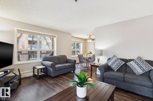 This living area features hardwood style flooring, a large window with a view of exterior buildings, and a dining area visible in the background - 202 17511 98A Avenue, Edmonton, AB - Indoor Photo Showing Living Room