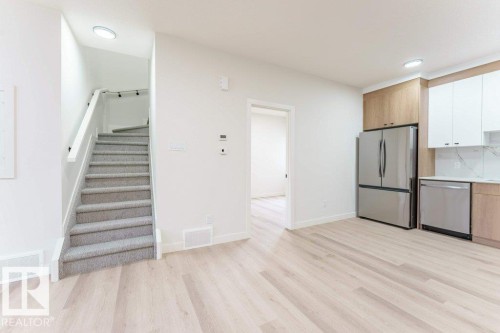Open-concept living area featuring light wood-finish flooring, a contemporary kitchen with stainless steel appliances, two-tone cabinetry, and white tile backsplash, and a staircase with light grey carpeting and a white handrail - 6920/6922 106 Street, Edmonton, AB - Indoor Photo Showing Kitchen
