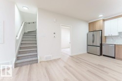 Open-concept living area featuring light wood-finish flooring, a contemporary kitchen with stainless steel appliances, two-tone cabinetry, and white tile backsplash, and a staircase with light grey carpeting and a white handrail - 