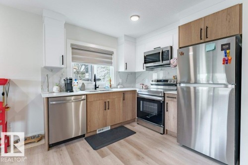 Modern kitchen featuring stainless steel appliances, a window with a roller shade, light wood-finish flooring, white and wood-tone cabinetry, and a white countertop with a patterned backsplash - 6920/6922 106 Street, Edmonton, AB - Indoor Photo Showing Kitchen