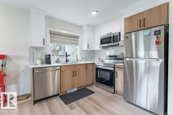 Modern kitchen featuring stainless steel appliances, a window with a roller shade, light wood-finish flooring, white and wood-tone cabinetry, and a white countertop with a patterned backsplash - 