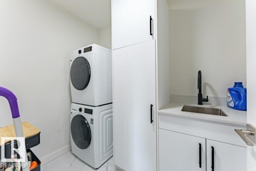 Laundry area featuring a stacked washer and dryer, white cabinetry with black hardware, a stainless steel sink with a matte black faucet, and a light-colored countertop - 6920/6922 106 Street, Edmonton, AB - Indoor Photo Showing Laundry Room