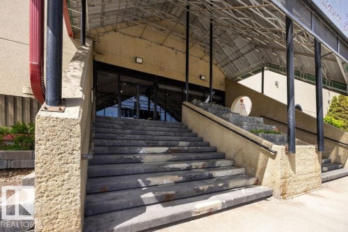 Concrete entryway featuring multiple sets of stairs, black metal railings, and a transparent overhead canopy - 1208 9918 101 Street, Edmonton, AB - 
