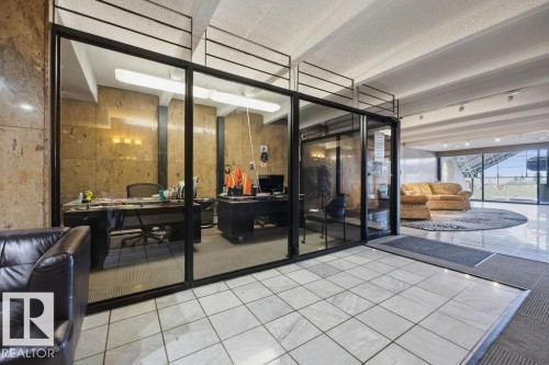 Entrance area featuring white square tile flooring transitioning to carpeted sections, with a glass-enclosed office space - 1208 9918 101 Street, Edmonton, AB - Indoor Photo Showing Other Room