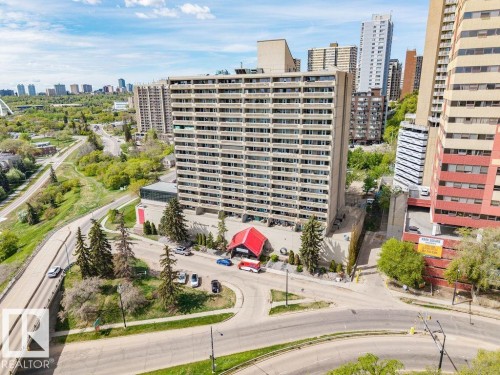 Multi-story building featuring numerous private balconies, a ground-level entrance with a red canopy, surrounding mature evergreen trees, and a developed road network - 1208 9918 101 Street, Edmonton, AB - Outdoor With Facade