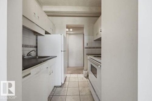 Galley kitchen featuring white cabinetry, a built-in dishwasher, white tile flooring, a dark countertop, and tile backsplash with a decorative border - 1208 9918 101 Street, Edmonton, AB - Indoor Photo Showing Kitchen