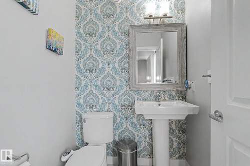Pedestal sink with chrome faucet, framed mirror, and decorative wallpaper accent wall, complemented by a white toilet and overhead vanity lighting - 18144 76 Street, Edmonton, AB - Indoor Photo Showing Bathroom