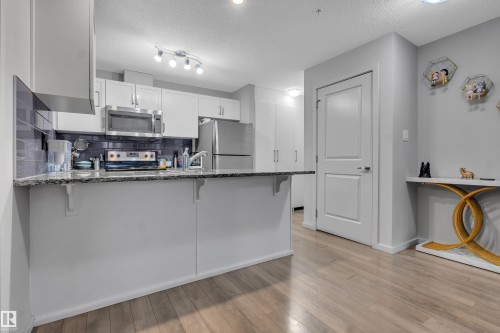 Kitchen featuring light wood-finish flooring, white cabinetry, a dark tiled backsplash, stainless steel appliances, and a peninsula with a stone-look countertop - 128 390 Windermere Road, Edmonton, AB - Indoor Photo Showing Kitchen