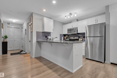 Open-concept kitchen featuring white cabinetry, stainless steel appliances, a breakfast bar, and wood-finish flooring - 128 390 Windermere Road, Edmonton, AB - Indoor Photo Showing Kitchen