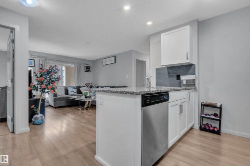 Open-concept kitchen island featuring a stainless steel dishwasher, white cabinetry, granite-style countertop, and a gray subway tile backsplash - 128 390 Windermere Road, Edmonton, AB - Indoor Photo Showing Kitchen