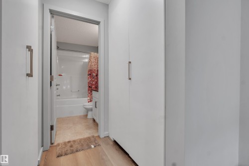 Hallway with wood-finish flooring featuring a built-in storage unit with contemporary hardware and a full bathroom entry - 128 390 Windermere Road, Edmonton, AB - Indoor