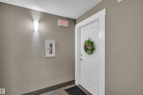 Entry corridor featuring a white panel door with modern hardware, textured wall finish, wood-finish flooring, and a contemporary wall sconce - 128 390 Windermere Road, Edmonton, AB - Indoor Photo Showing Other Room