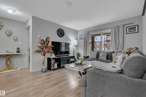 Bright living space featuring wood-finish flooring, light gray walls, and an expansive window with drapes - 128 390 Windermere Road, Edmonton, AB - Indoor Photo Showing Living Room