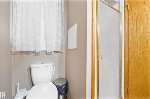 Compact bathroom featuring a white toilet, window with privacy curtain, and a shower with a frosted glass door and brass-tone framing - 4231 32 Avenue, Edmonton, AB - Indoor Photo Showing Bathroom