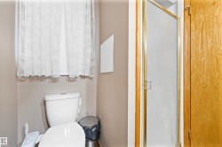 Compact bathroom featuring a white toilet, window with privacy curtain, and a shower with a frosted glass door and brass-tone framing - 