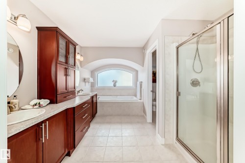 Spacious bathroom featuring a double vanity with cherry wood-finish cabinetry, a large built-in soaking tub under an arched window, a separate glass-enclosed shower, and light-toned tile flooring - 1330 114B Street, Edmonton, AB - Indoor Photo Showing Bathroom