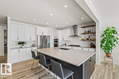 Spacious kitchen featuring an expansive island with a light-toned countertop, white cabinetry with contemporary hardware, stainless steel appliances, a subway tile backsplash, and wood-finish flooring - 21003 131 Avenue, Edmonton, AB 