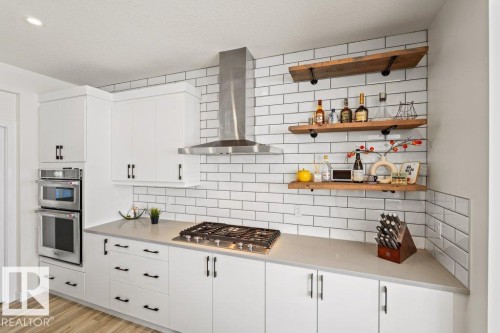 Modern kitchen featuring white cabinetry, light-toned countertops, a gas cooktop with stainless steel range hood, and built-in double ovens - 21003 131 Avenue, Edmonton, AB 