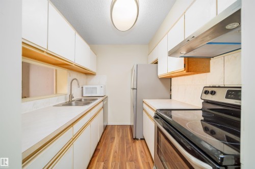 Galley kitchen featuring white cabinetry, light countertops, wood-finish flooring, a stainless steel double basin sink, and a built-in range hood - 213 5065 31 Avenue, Edmonton, AB - Indoor Photo Showing Kitchen With Double Sink