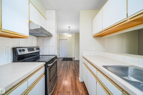 Galley kitchen featuring white cabinetry with wood-finish trim, a stainless steel range hood, and a built-in stainless steel double sink - 213 5065 31 Avenue, Edmonton, AB - Indoor Photo Showing Kitchen