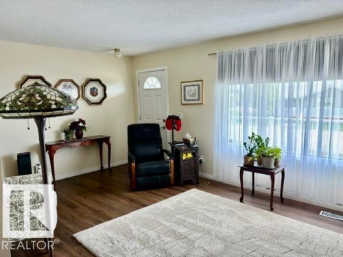Spacious living area featuring wood-finish flooring, a prominent window with sheer curtains, neutral wall paint, and a white entry door with an arched glass insert - 4805 53Ave, Willingdon, AB - Indoor