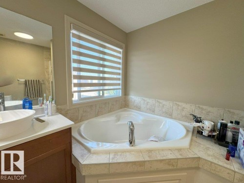 Corner soaking tub with tile surround, a large window with patterned blinds, and a vanity with a vessel sink and mirror - 1206 Mckinney Court, Edmonton, AB - Indoor Photo Showing Bathroom