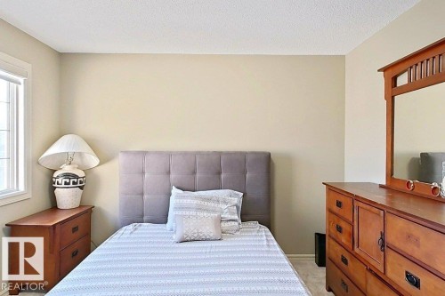 Neutral-toned room featuring a window with white trim, textured ceiling, light-colored carpeting, and a visible portion of a wooden dresser with a mirror - 1206 Mckinney Court, Edmonton, AB - Indoor Photo Showing Bedroom