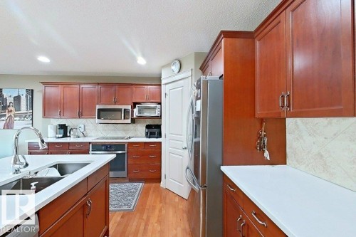 Kitchen featuring wood-finish flooring, cherry-toned cabinetry, stainless steel appliances, a center island with a double basin sink and gooseneck faucet, and white countertops - 1206 Mckinney Court, Edmonton, AB - Indoor Photo Showing Kitchen With Double Sink