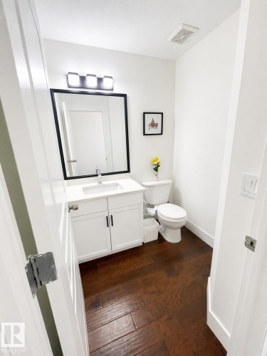 This bathroom features rich hardwood flooring, a white vanity with an integrated sink and storage, and a mirror with a dark frame, complemented by a modern three-light fixture - 3243 26 Street, Edmonton, AB - Indoor Photo Showing Bathroom