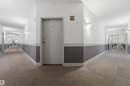 Well-lit hallway featuring a light gray door, patterned carpet flooring, white walls with a dark gray wainscoting, and modern wall-mounted sconces - 302 2508 50 Street Nw, Edmonton, AB - Indoor Photo Showing Other Room