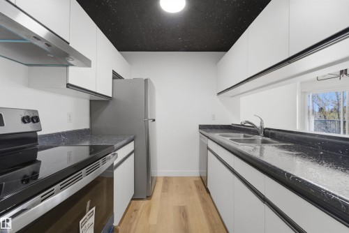 Galley kitchen featuring wood-finish flooring, white cabinetry, dark countertops, and stainless steel appliances including a range, refrigerator, and dual basin sink - 302 2508 50 Street Nw, Edmonton, AB - Indoor Photo Showing Kitchen With Double Sink