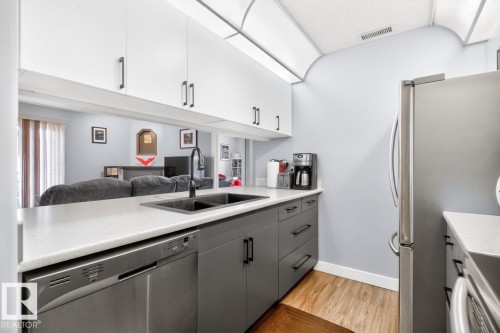 Kitchen with two-tone cabinetry featuring white upper cabinets and gray lower cabinets, light-toned countertops, a stainless steel dishwasher, a dark basin sink with a matte black faucet, and wood-finish flooring - 220 16303 95 Street, Edmonton, AB - Indoor Photo Showing Kitchen With Double Sink