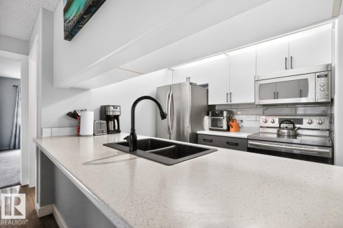 Kitchen featuring light-toned countertops, a matte black double basin sink with a matching gooseneck faucet, stainless steel appliances, and white upper cabinetry - 220 16303 95 Street, Edmonton, AB - Indoor Photo Showing Kitchen With Double Sink With Upgraded Kitchen