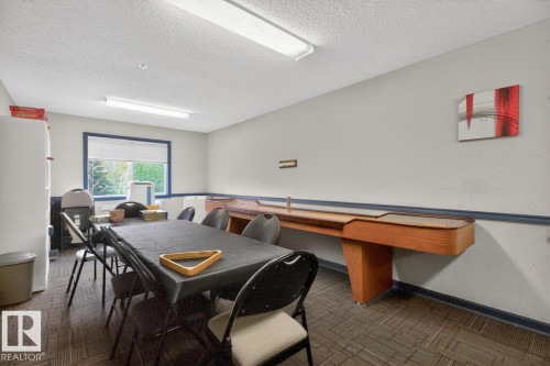 Recreational room featuring a shuffleboard table, a window with blinds, and patterned carpet flooring - 220 16303 95 Street, Edmonton, AB - Indoor