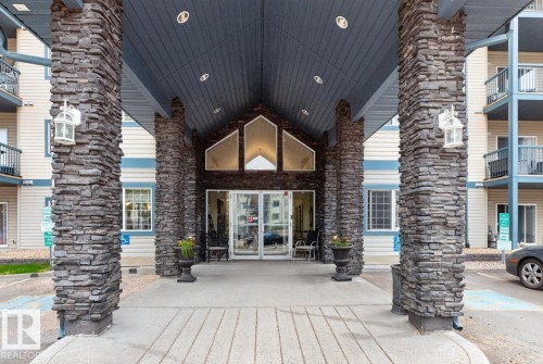 Building entrance featuring stacked stone columns, a covered portico with recessed lighting, and a gabled architectural detail above the glass entry doors - 220 16303 95 Street, Edmonton, AB - Outdoor With Balcony With Facade