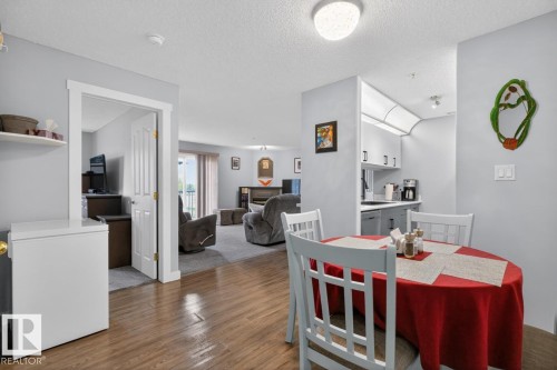 Open-concept dining area with wood-finish flooring, featuring a recessed kitchen with upper and lower cabinetry, and a carpeted living room beyond - 220 16303 95 Street, Edmonton, AB - Indoor Photo Showing Dining Room