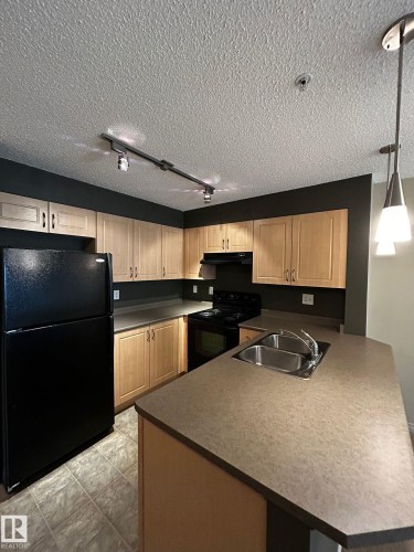 Kitchen featuring light wood-finish cabinetry, a double basin stainless steel sink, black appliances, and track lighting - 205 11445 Ellerslie Road, Edmonton, AB 