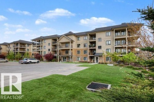 Multi-story building featuring light-toned siding, dark roofing, stone accents, multiple balconies, and an adjacent paved parking area - 209 1029 173 Stnw, Edmonton, AB - Outdoor With Balcony With Facade