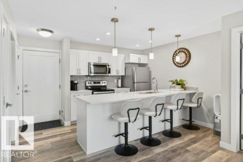 Kitchen featuring a white quartz-topped island with seating, stainless steel appliances, white cabinetry, and wood-finish flooring - 209 1029 173 Stnw, Edmonton, AB - Indoor Photo Showing Kitchen With Stainless Steel Kitchen With Upgraded Kitchen