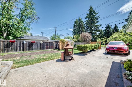 Paved concrete patio and driveway area featuring a rectangular planted garden bed, a wooden fence, mature trees, and a visible portion of a green gutter system - 11309 115 Street, Edmonton, AB - Outdoor