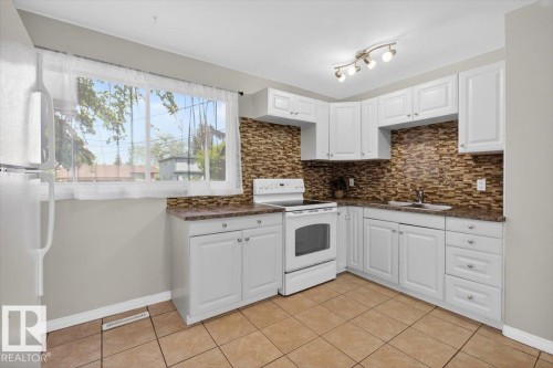 Bright kitchen featuring white cabinetry, a stone-finish backsplash, and durable tile flooring - 4630 114 Avenue, Edmonton, AB - Indoor Photo Showing Kitchen With Double Sink