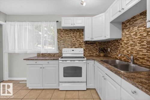 Kitchen featuring white cabinetry, a mosaic tile backsplash, a double basin sink, a white electric range, and light tile flooring - 4630 114 Avenue, Edmonton, AB - Indoor Photo Showing Kitchen With Double Sink
