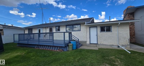 Rear exterior featuring an elevated deck with glass and wood railing, stucco siding, and a maintained lawn - 5113 45 B Avenue, Vegreville, AB - Outdoor With Deck Patio Veranda