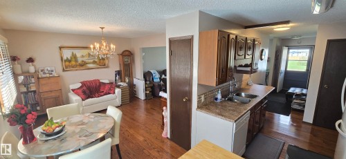 Kitchen with wood-finish cabinetry, double basin stainless steel sink, and dishwasher, offering sightlines into the dining area with wood-finish flooring and a chandelier - 5113 45 B Avenue, Vegreville, AB - Indoor