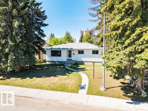 White single-story residence with a gray shingle roof, front lawn, mature evergreen trees, and a curved concrete walkway - 8714 164 Street, Edmonton, AB 